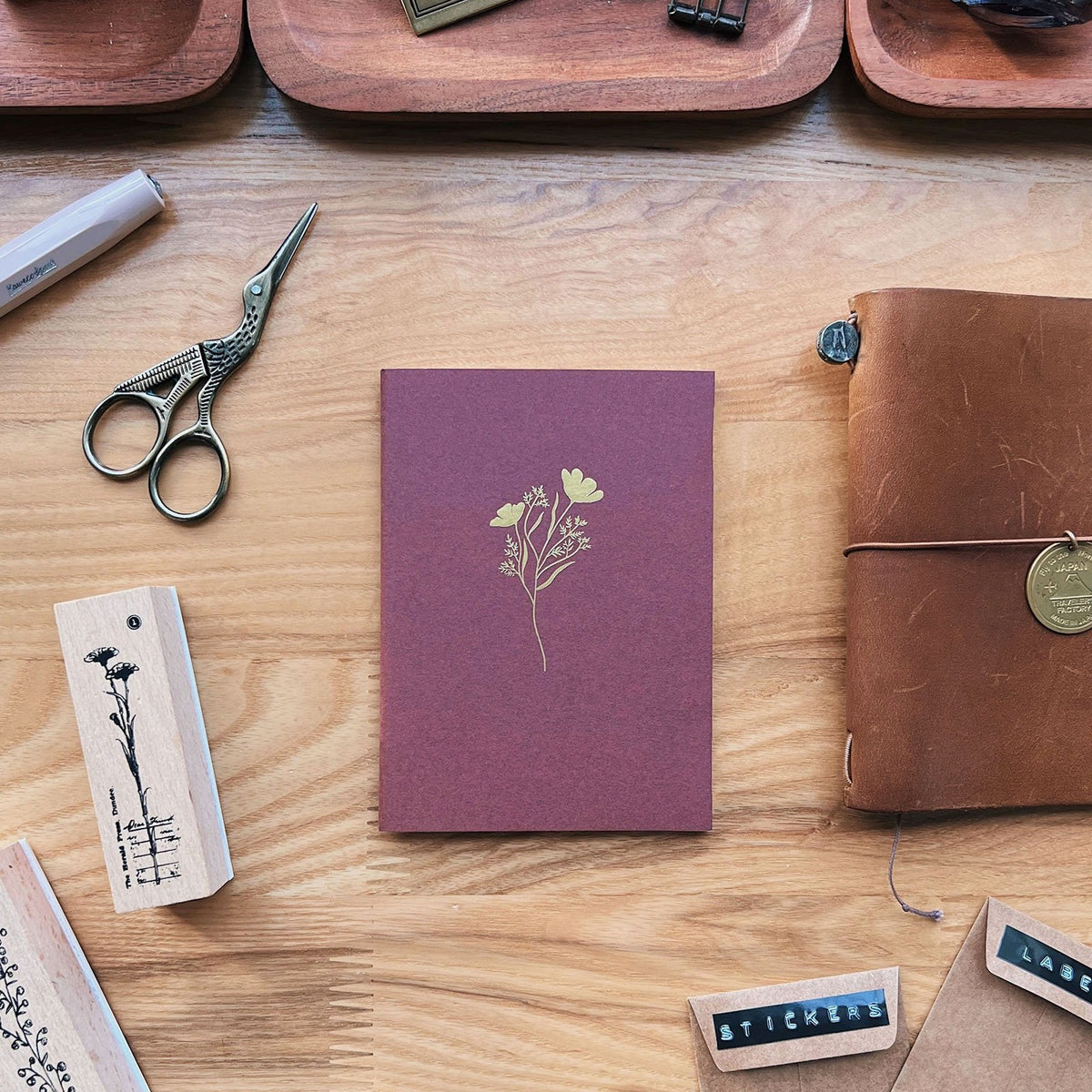 Sarica Studio Floral Notebook, passport TN size, bordeaux with a gold floral design, beside rubber stamps and a leather traveler's notebook