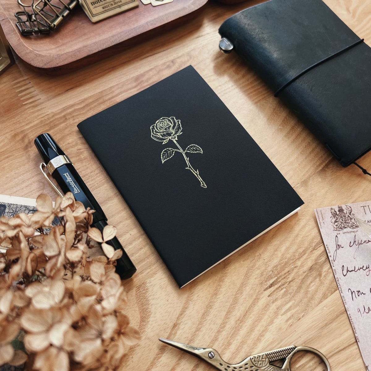 Sarica Studio Rose Notebook, passport TN size, black with a gold rose design, beside a black leather traveler's notebook