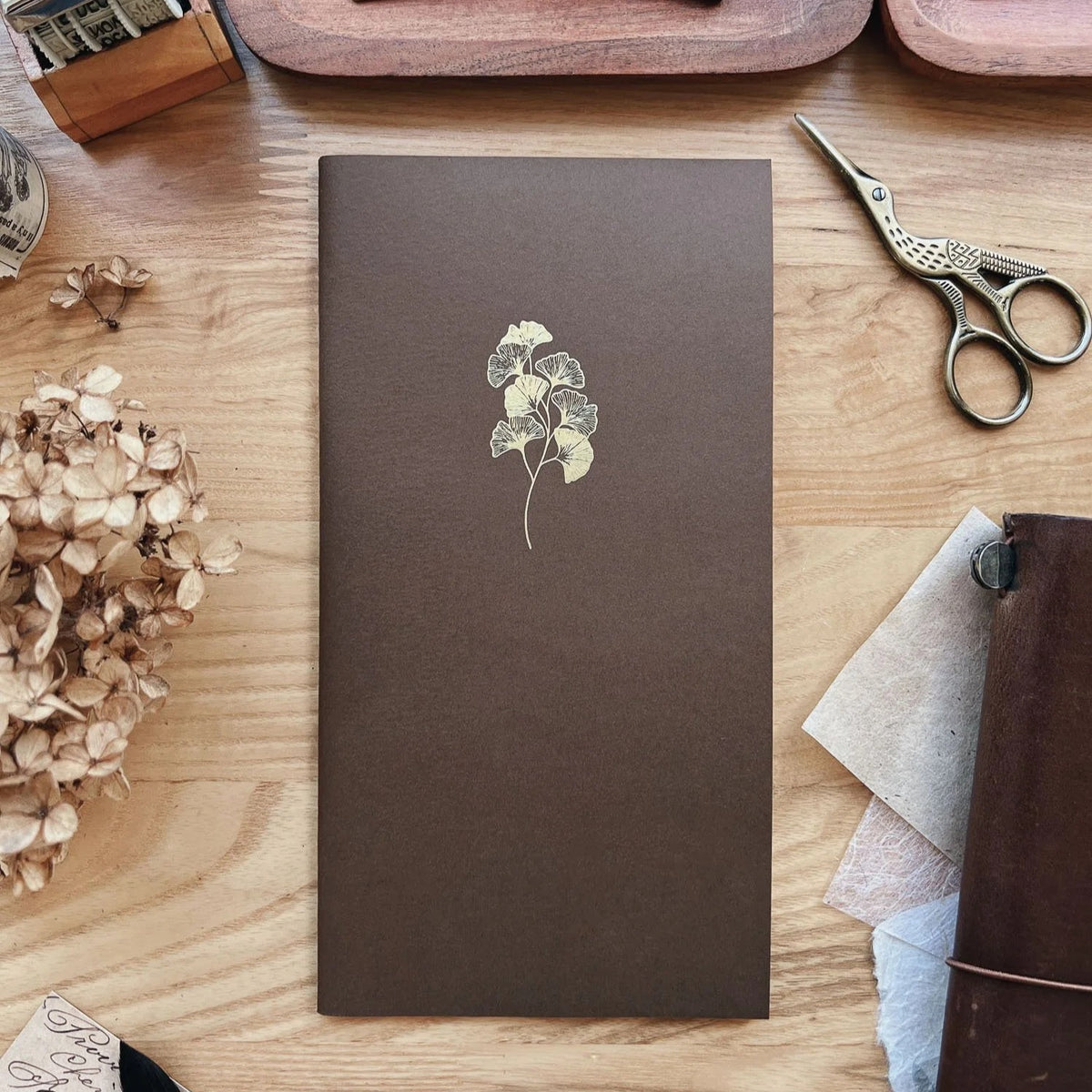 Sarica Studio Ginkgo Notebook, standard TN size, chestnut brown cover with a gold debossed ginkgo design, beside dried hydrangeas