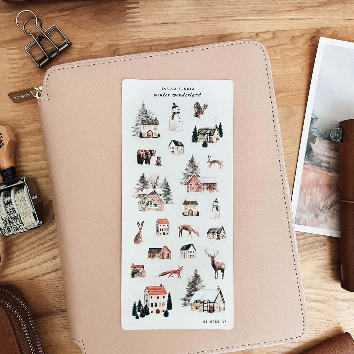 Sarica Studio Winter Wonderland sticker sheet of snowy houses and woodland animals on a blush leather notebook cover